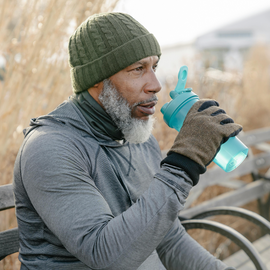 A bearded man wearing gloves drinks from a water bottle.